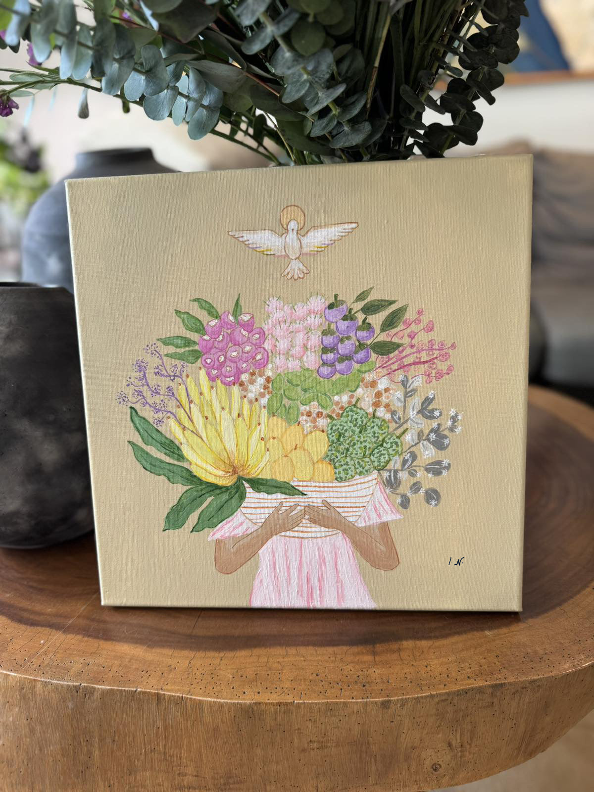 A painting titled 'Fruits of the Holy Spirit' by Ica Narvaez, featuring a floral arrangement with various colored flowers and fruits on a wooden surface, placed in a home environment with a vase of purple flowers in the background.