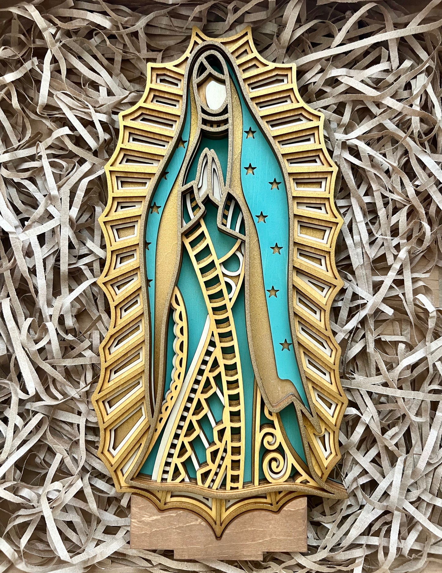 A piece of wood art depicting Our Lady of Guadalupe with a combination of mandala art and 20th century art deco designs, placed on a shredded brown paper.