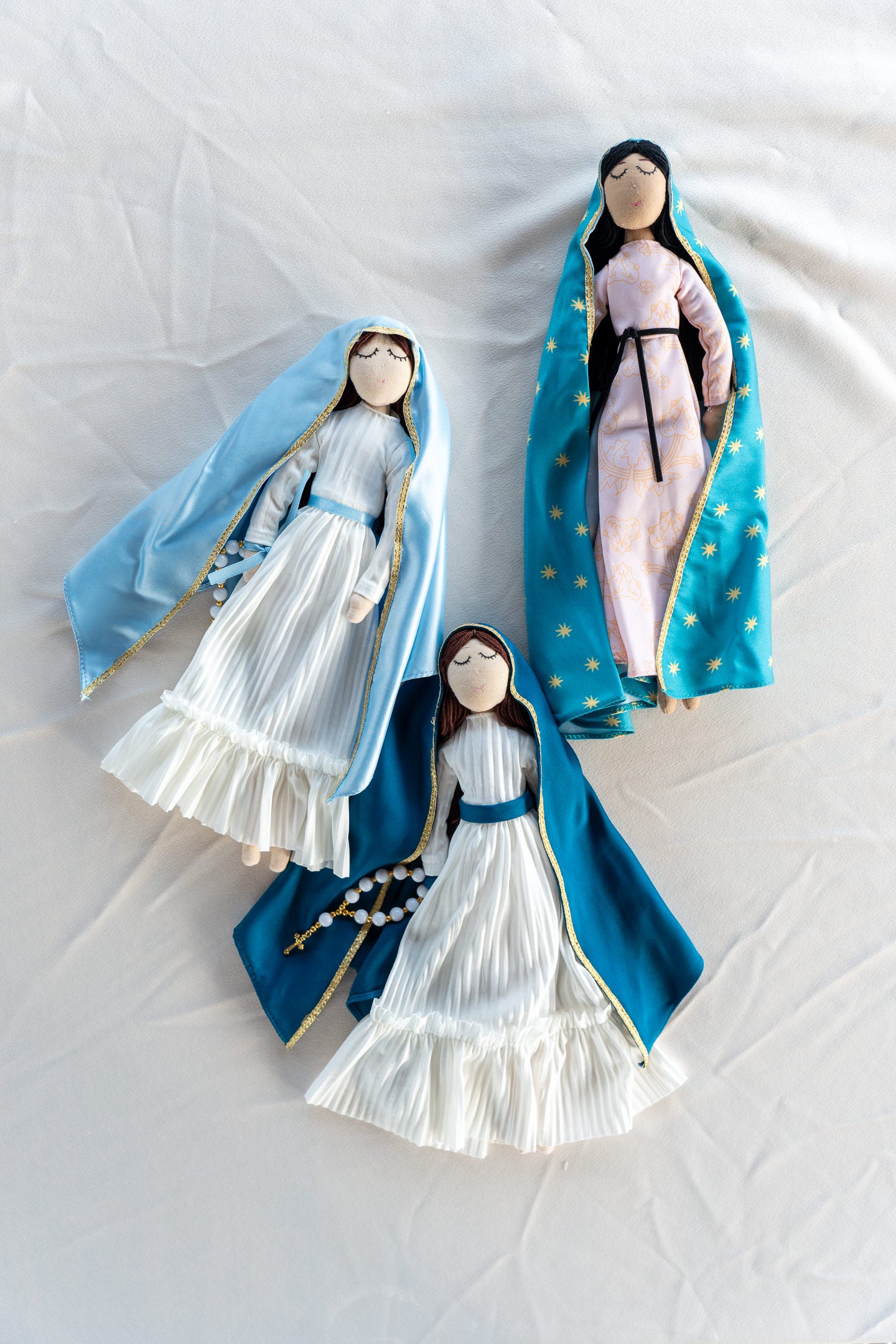 Three handmade dolls, depicting a young girl in a blue and white dress, and the other a woman in a white and blue veil, both with intricate details and accessories.