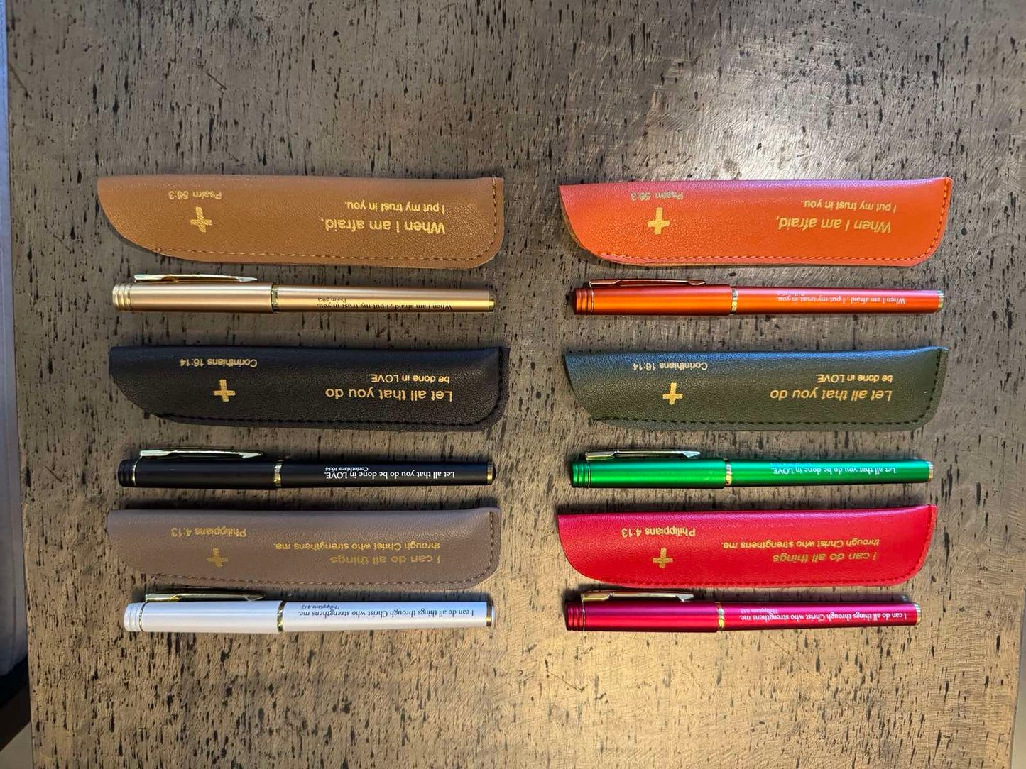A collection of colored and neutral pens with leather sleeves, each inscribed with an inspirational Bible quote.
