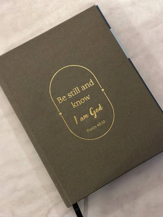 A gray journal with a premium linen material book cover, featuring a quote from Psalm 46:10 'Be still and know I am God' in gold lettering.
