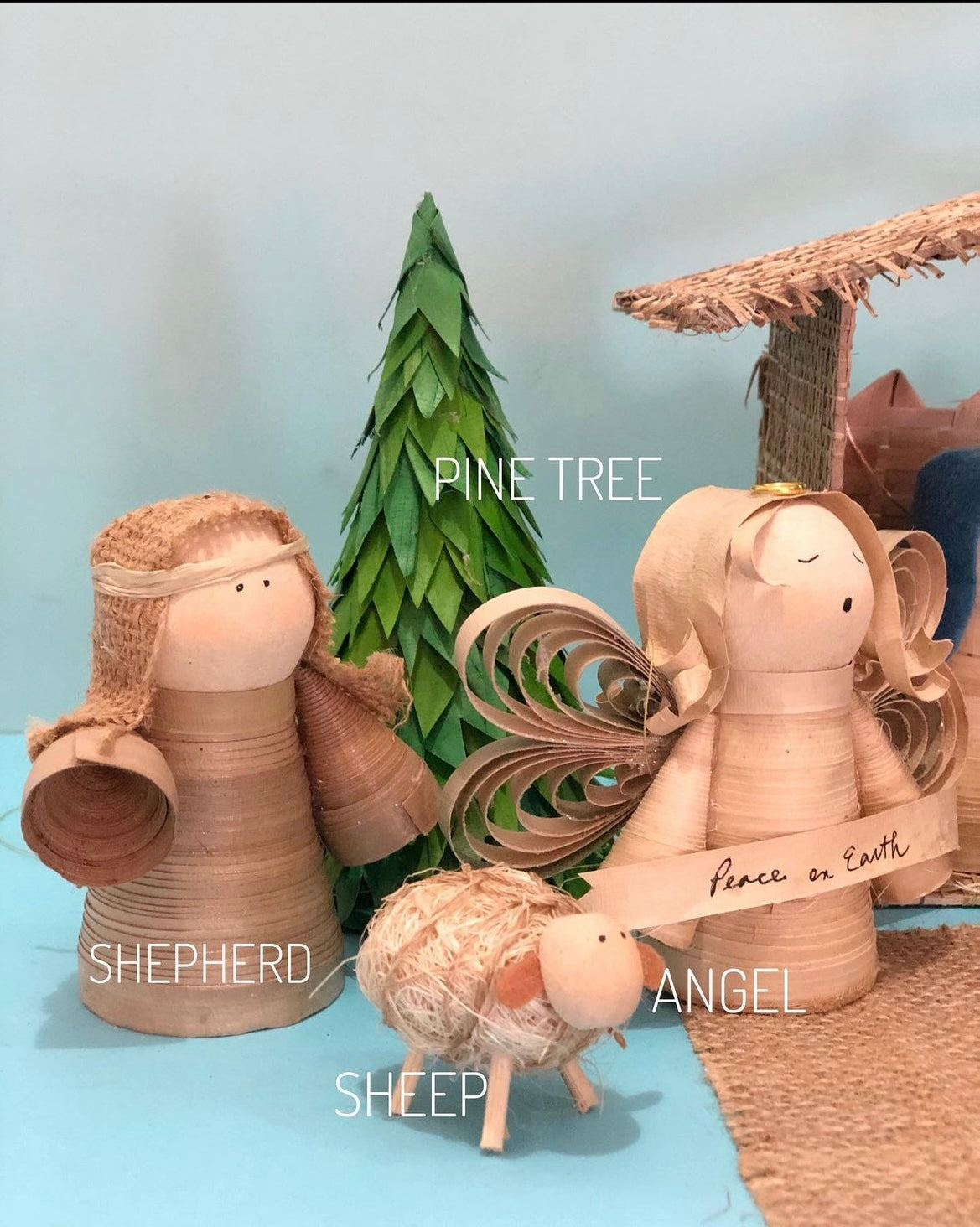 A wooden nativity set featuring various figures including the shepherd, angel and a sheep, with a detached roof and wall, a pine tree, and a burlap mat.