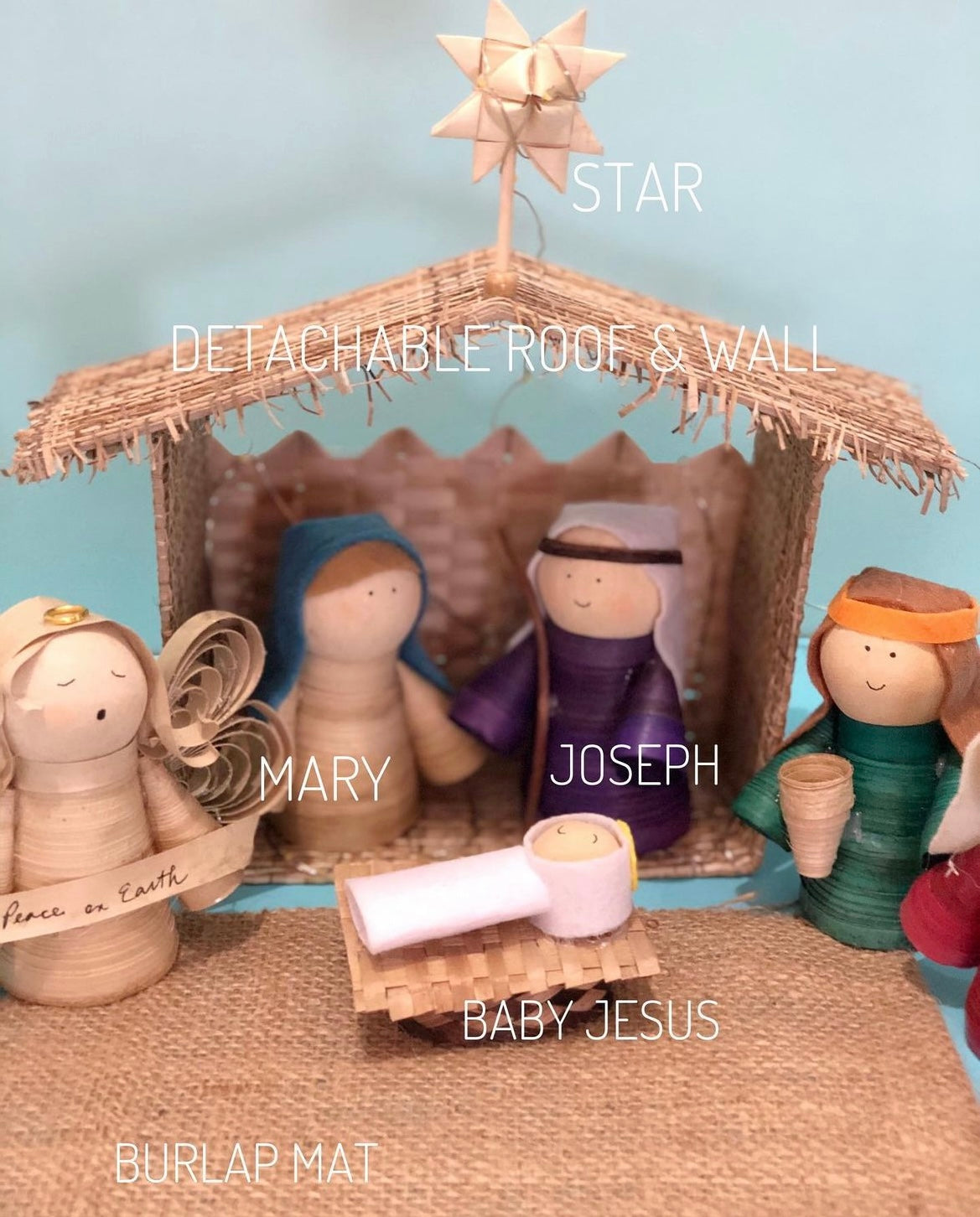A close up image of a wooden nativity set featuring various figures including the baby Jesus, Mary, Joseph, the three Wise Men, and the angel with a detached roof and wall, a pine tree, and a burlap mat.