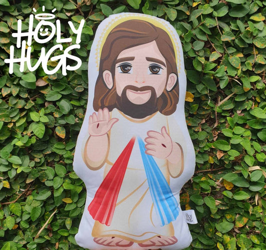 A beige-colored pillow with an illustration of a smiling figure of Jesus, with a blue, red, and white sash. The pillow has the text 'Holy Hugs' on it.