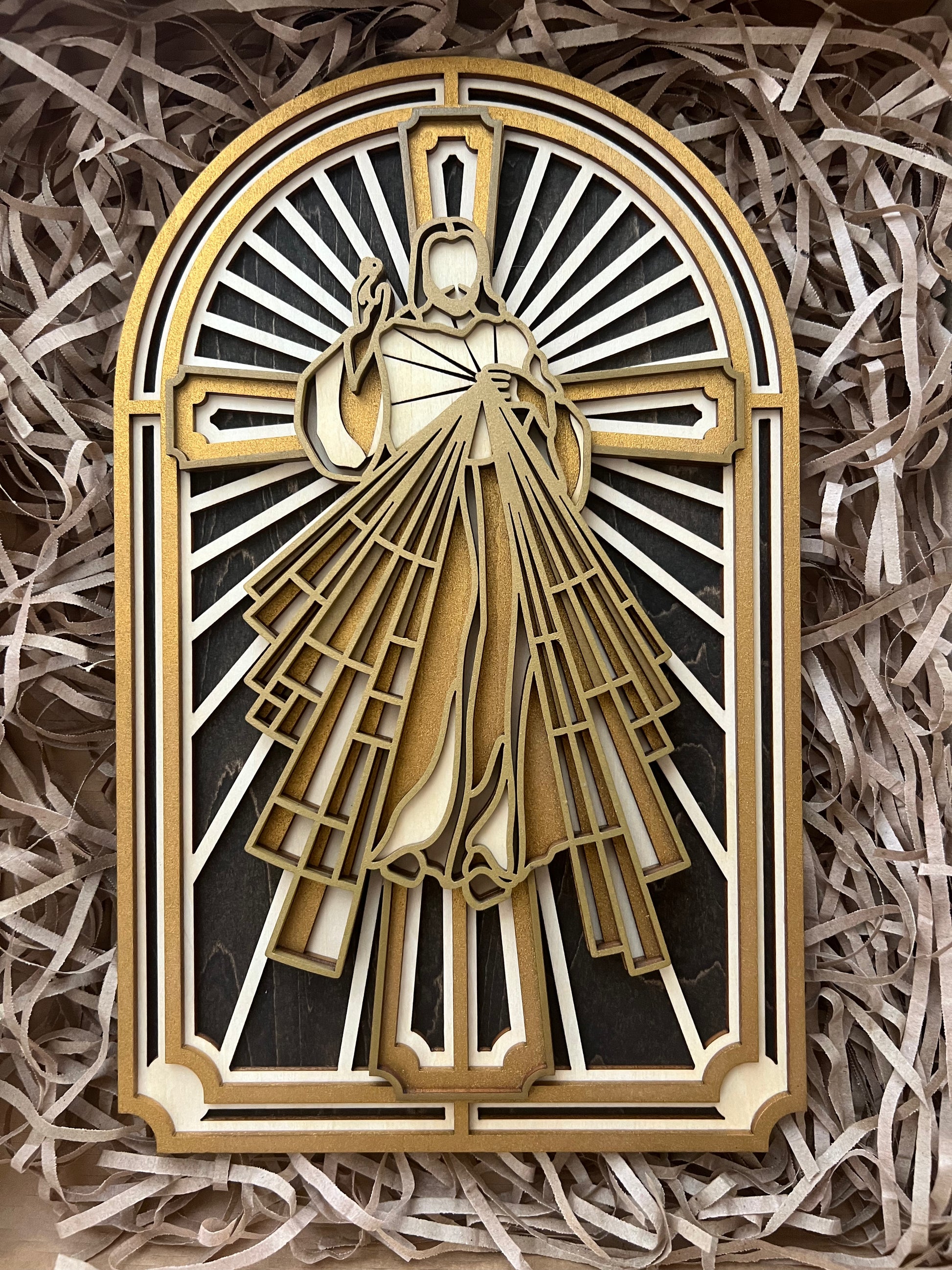 A multi-layered wood art depiction of the Sacred Heart of Jesus Christ, with a gold and white color scheme, placed on a shredded brown paper. 