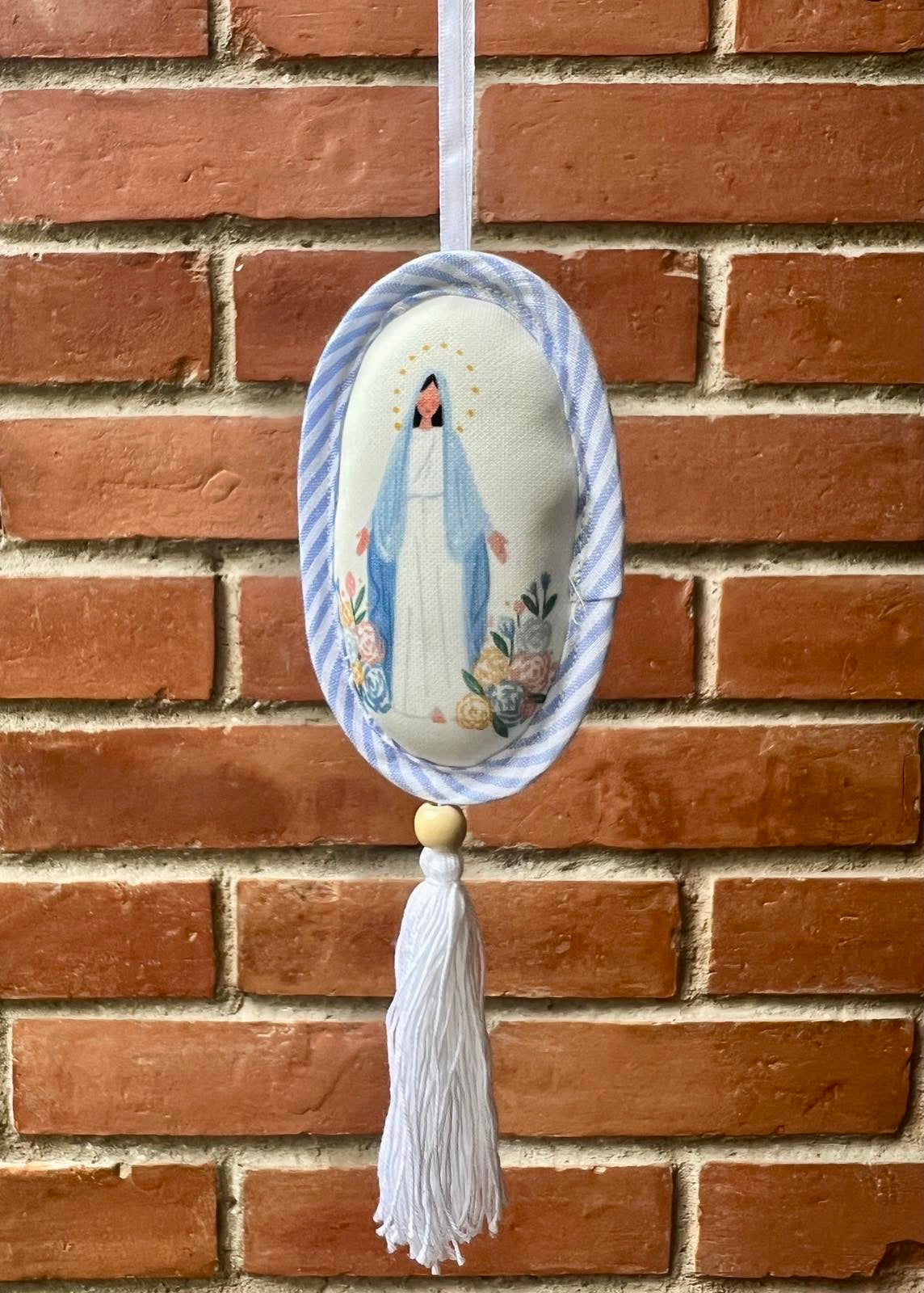 Decorative oval shaped ornament depicting Our Blessed Mother in a blue striped border and a white tassel against a brick wall.