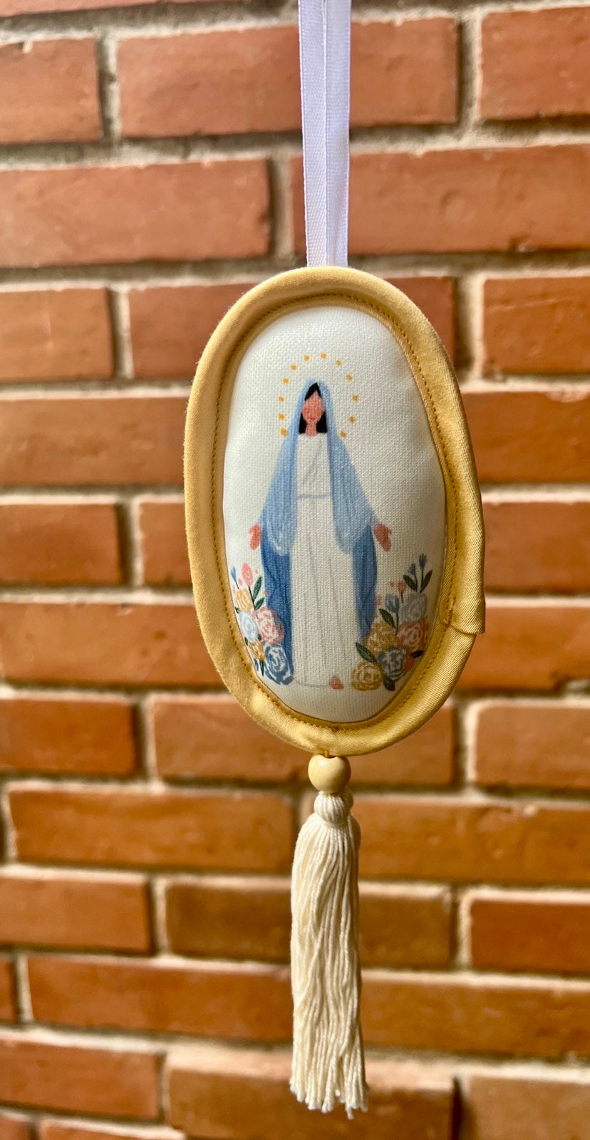 Decorative oval shaped ornament depicting Our Blessed Mother in a yellow  border and a cream tassel against a brick wall.