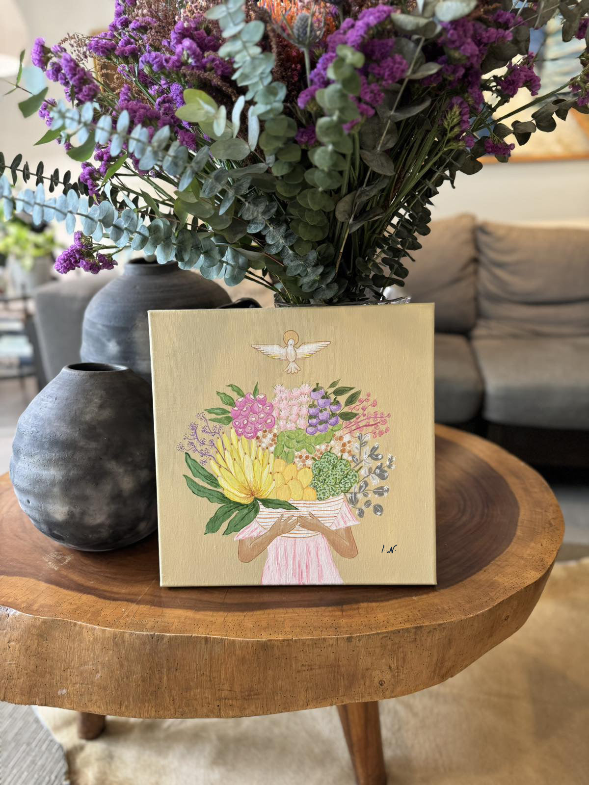 A painting titled 'Fruits of the Holy Spirit' by Ica Narvaez, featuring a floral arrangement with various colored flowers and fruits on a wooden surface, placed in a home environment with a vase of purple flowers in the background.