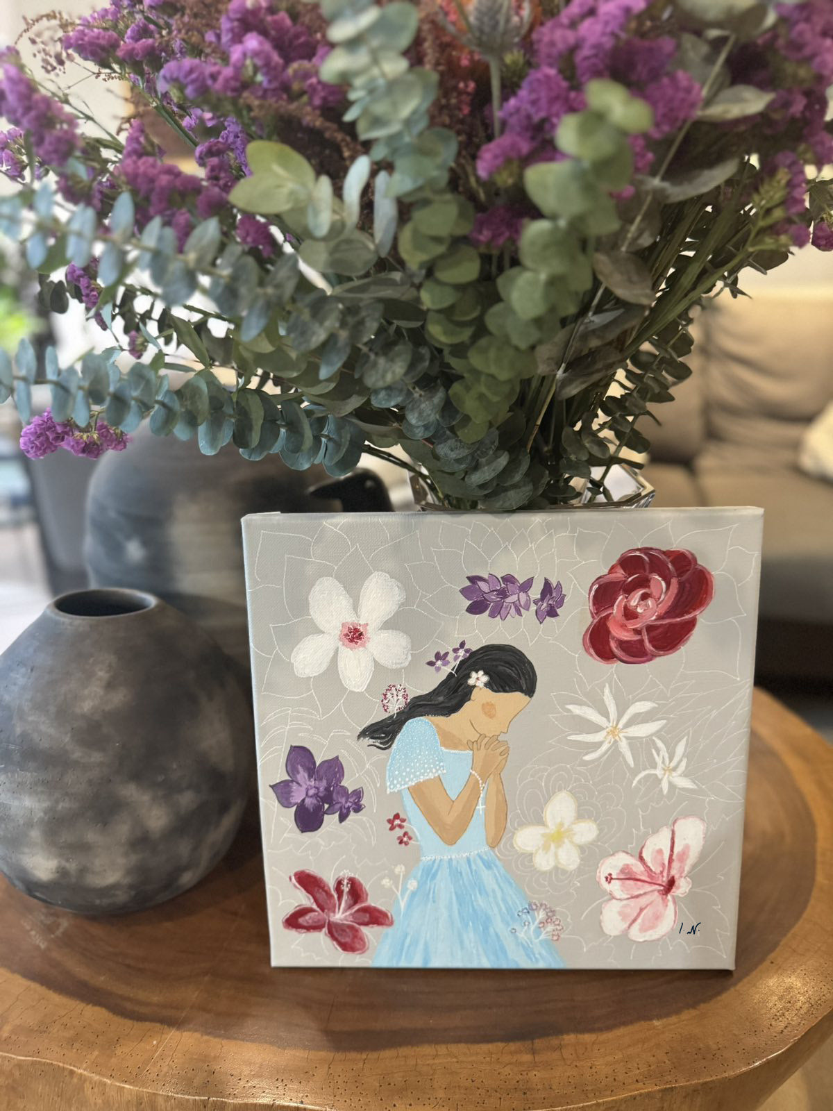 A painting depicting a girl with a floral background, surrounded by various flowers and a symbolic representation of the rosary, placed on a wooden surface with a plant in the background.