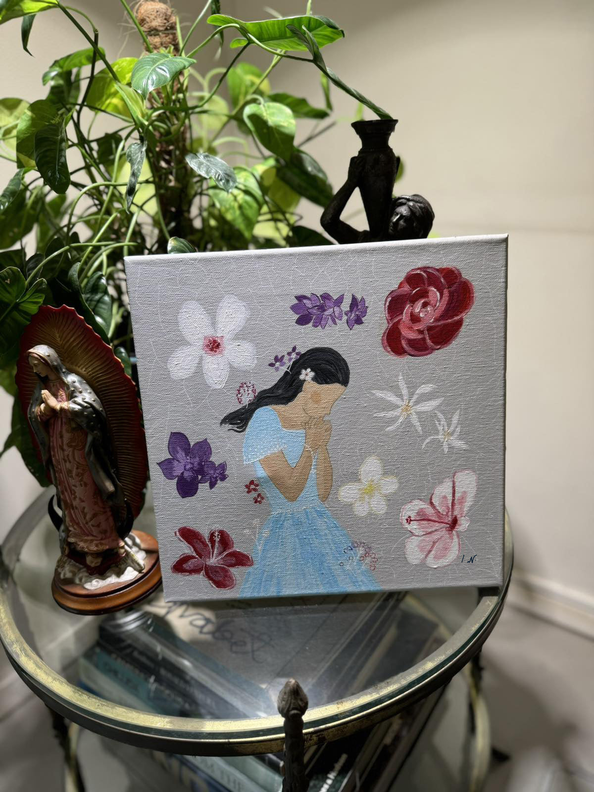 A painting depicting a girl with a floral background, surrounded by various flowers and a symbolic representation of the rosary, placed on a glass surface with a plant and a statue in the background.