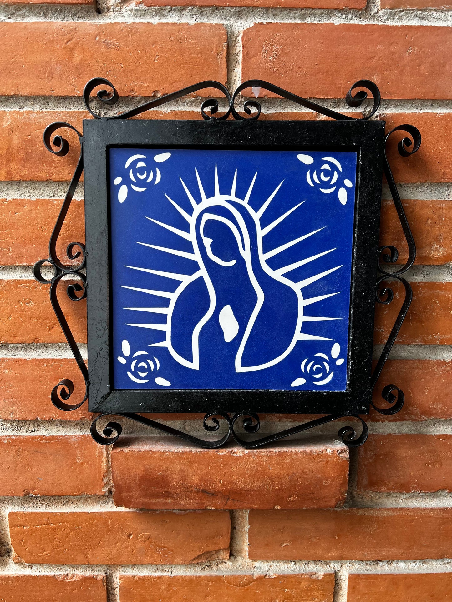 A blue decorative malaga tiles with a white floral design and feature of Our Lady of Guadalupe on a wrought-iron frame, placed on a brick wall.