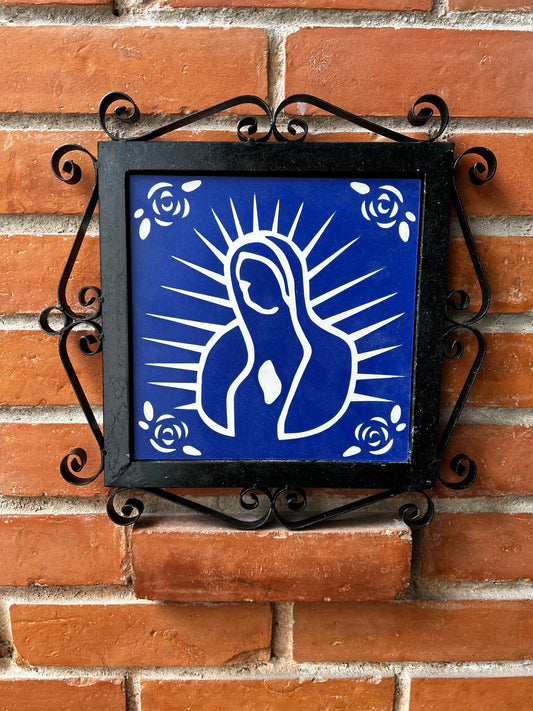 A blue decorative malaga tiles with a white floral design and feature of Our Lady of Guadalupe on a wrought-iron frame, placed on a brick wall.
