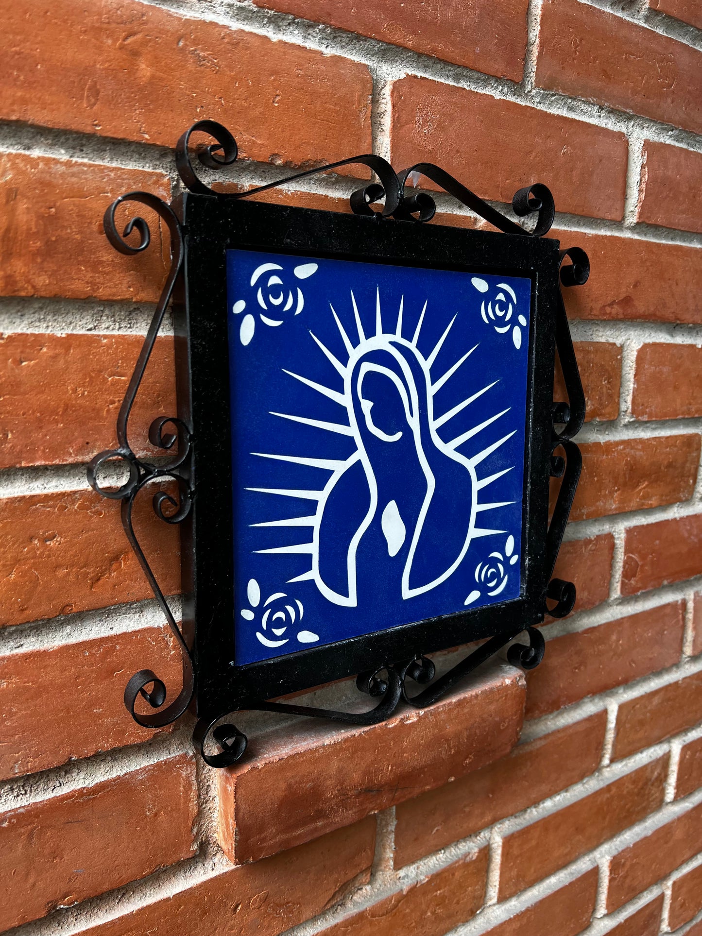 A blue decorative malaga tiles with a white floral design and feature of Our Lady of Guadalupe on a wrought-iron frame, placed on a brick wall.