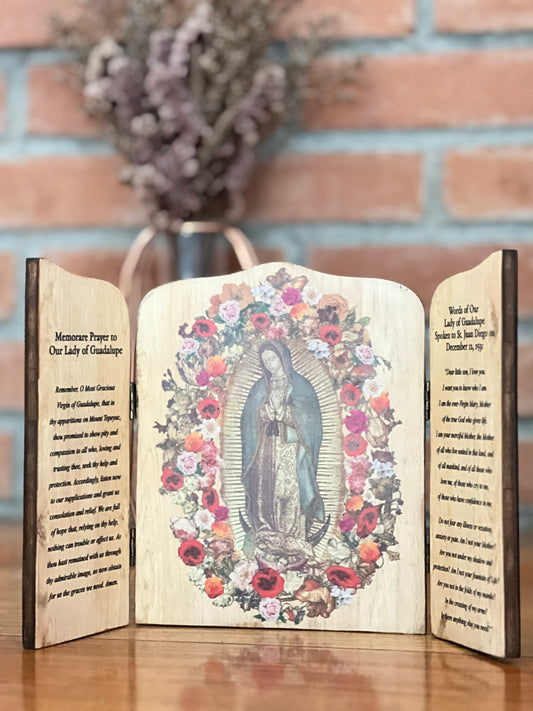 A wooden triptych featuring an image of Our Lady  of Guadalupe with floral designs, with inscriptions on the shutters. The left shutter is inscribed with a prayer, while the right shutter has the words of Our Lady to St. Juan Diego.