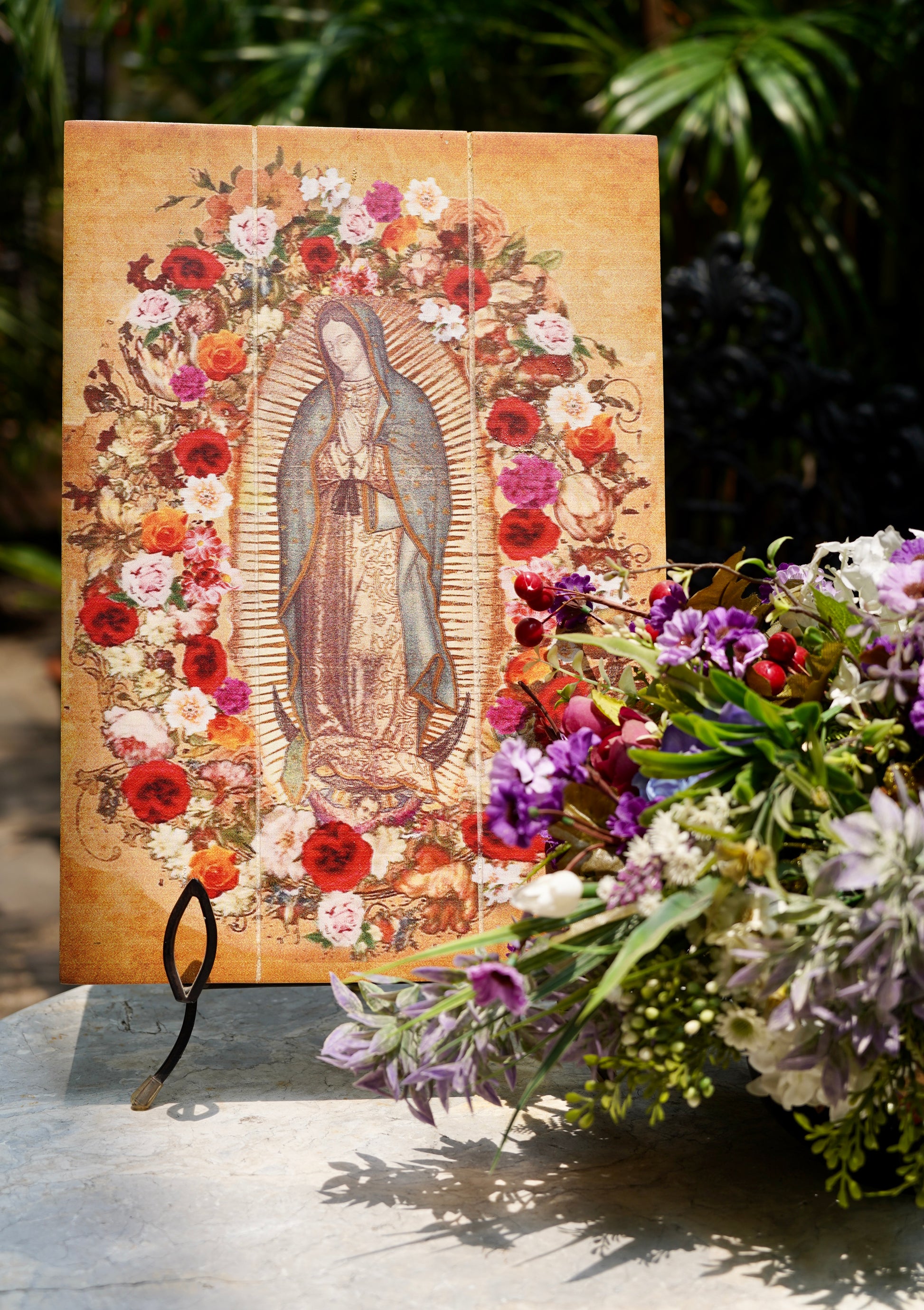 A tablet with an image of Our Lady  of Guadalupe surrounded by floral patterns, displayed outdoors with a stand not visible in the image.