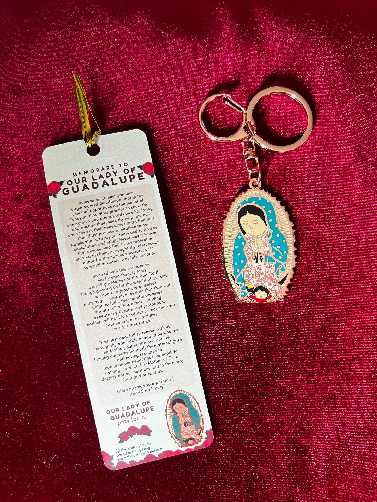 A key chain featuring a bronze-colored Our Lady of Guadalupe together with a bookmark with a prayer and rose designs, placed on a red surface.
