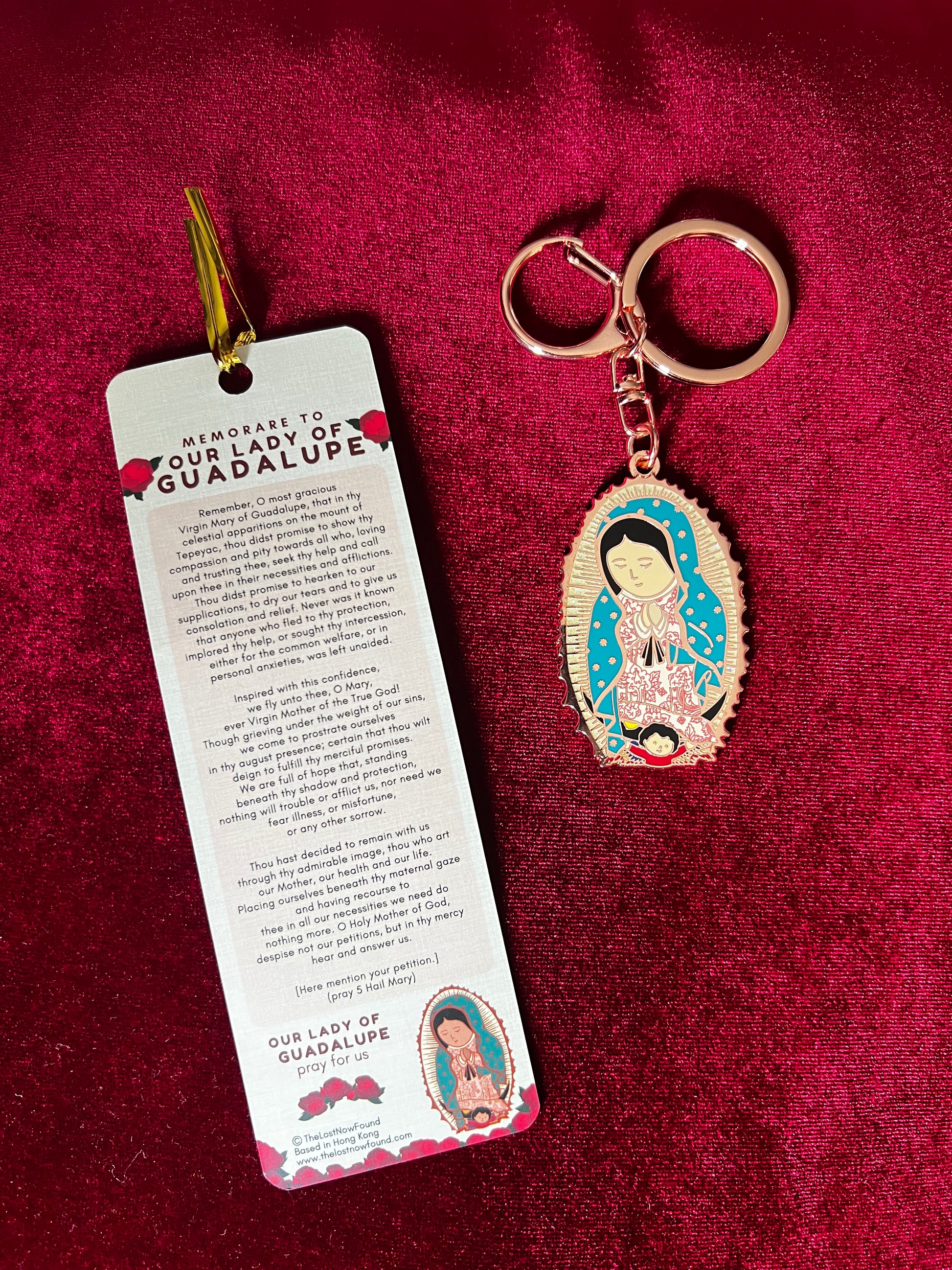 A key chain featuring a bronze-colored Our Lady of Guadalupe together with a bookmark with a prayer and rose designs, placed on a red surface.