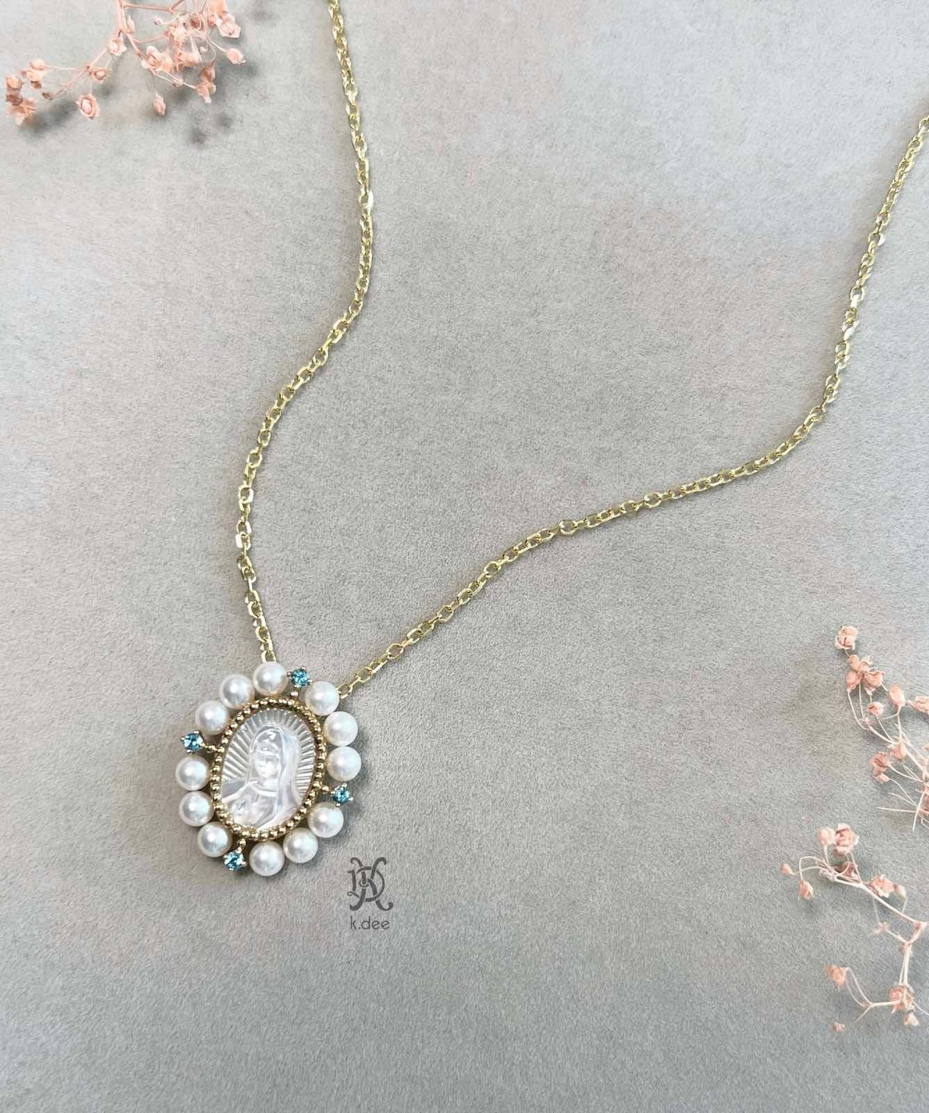 A gold necklace with a pendant featuring a hand-carved mother-of-pearl portrait of Our Lady of Guadalupe, surrounded by blue topaz stones and fresh water pearls.