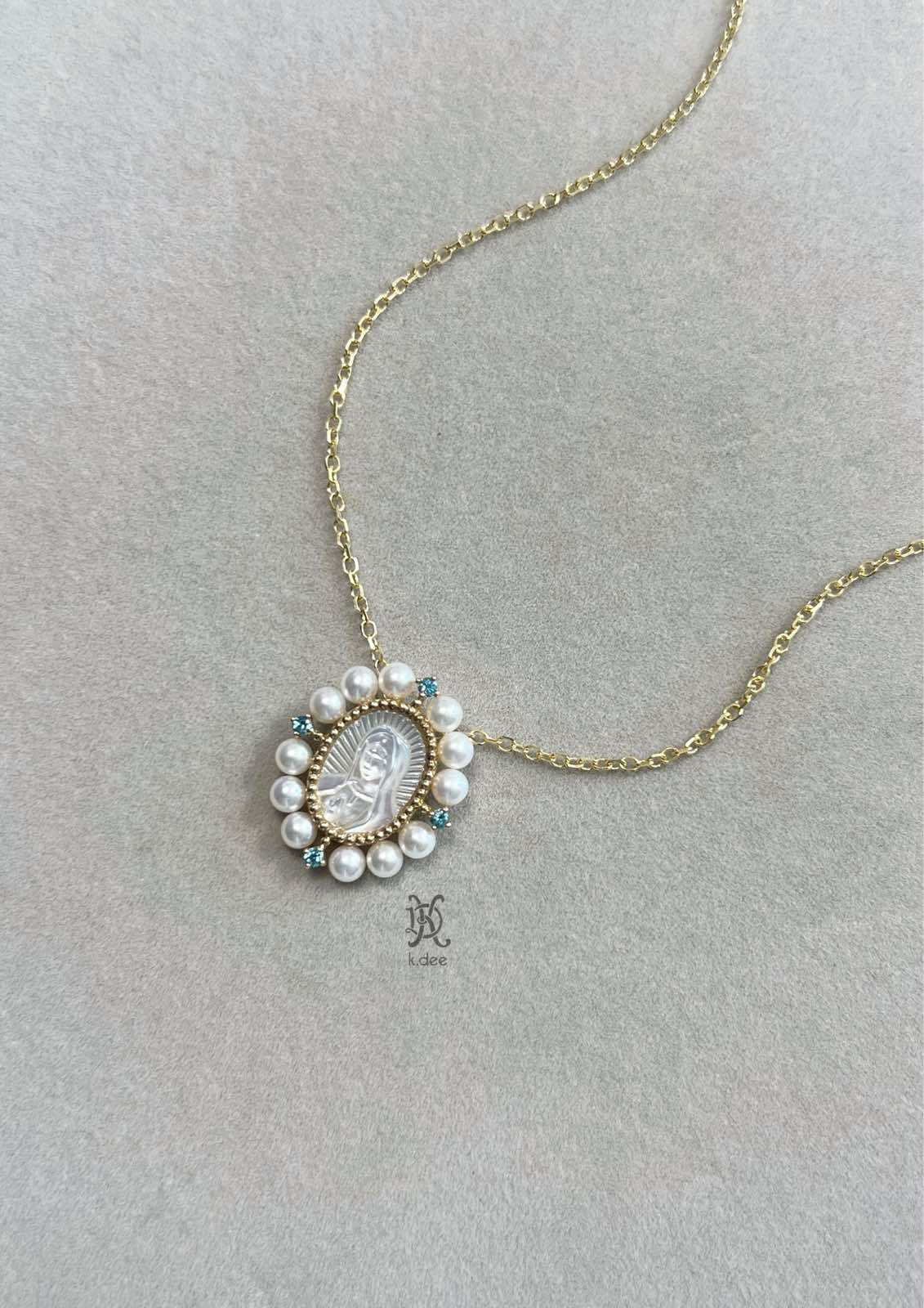 A gold necklace with a pendant featuring a hand-carved mother-of-pearl portrait of Our Lady of Guadalupe, surrounded by blue topaz stones and fresh water pearls.