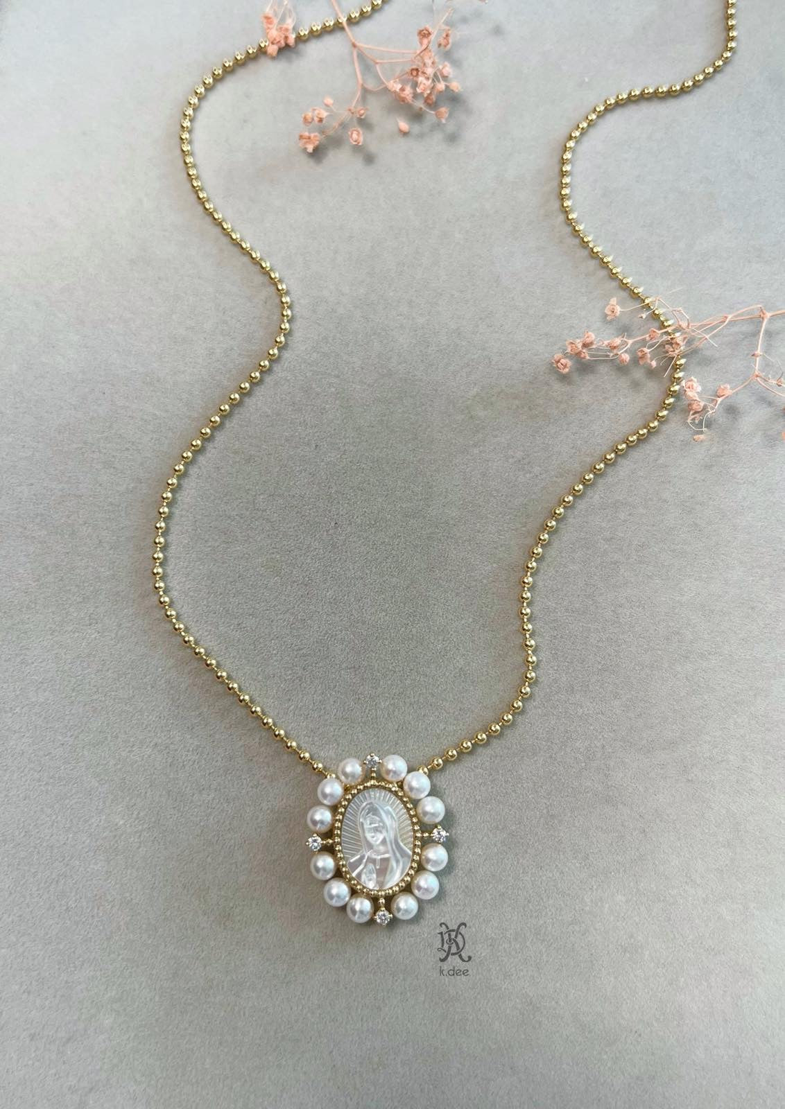 A 1-inch oval pendant of Our Lady of Guadalupe, hand-carved from mother-of-pearl, in 14K yellow gold, displayed on a gold chain.