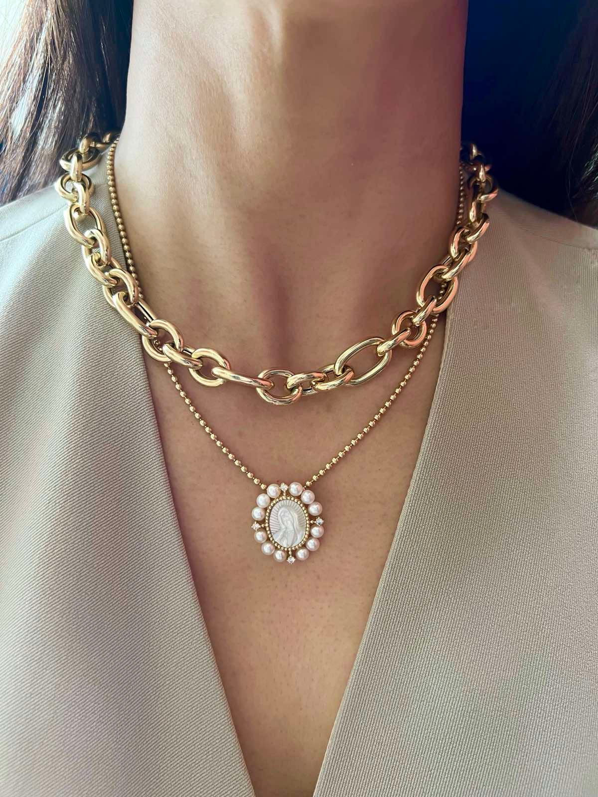 A 1-inch oval pendant of Our Lady of Guadalupe, hand-carved from mother-of-pearl, in 14K yellow gold, displayed on a gold chain. 