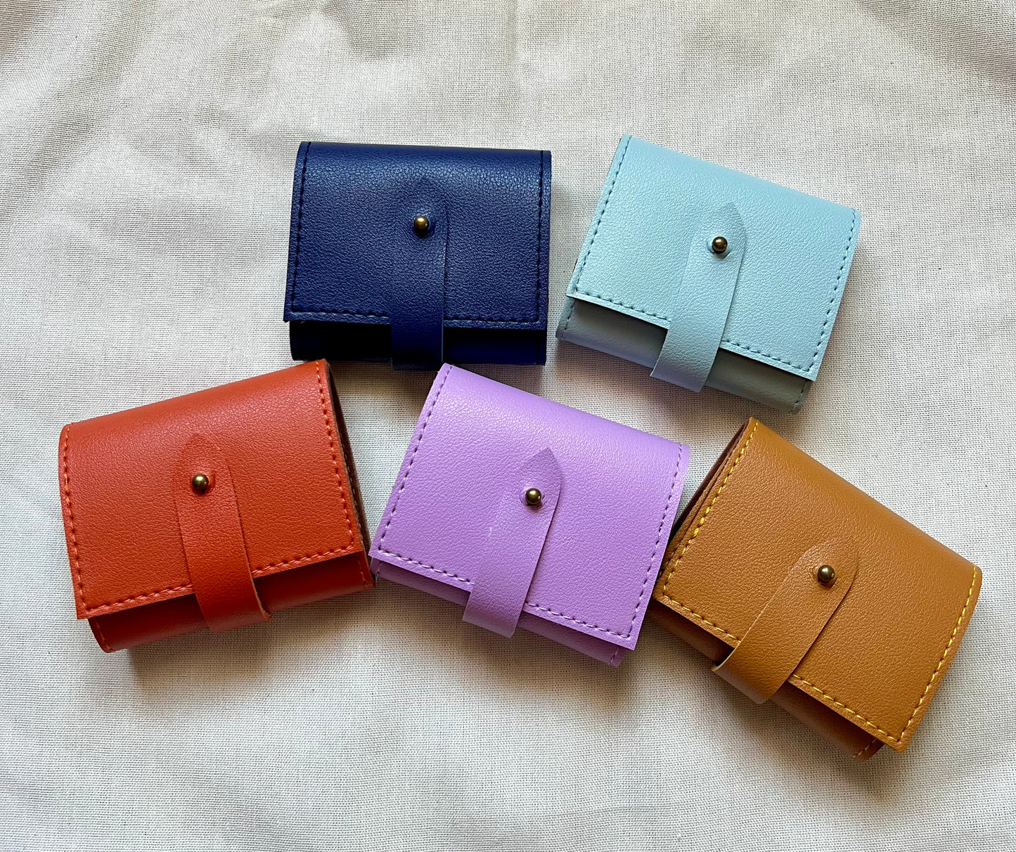 Five small leather wallets in different colors arranged on a light fabric background.