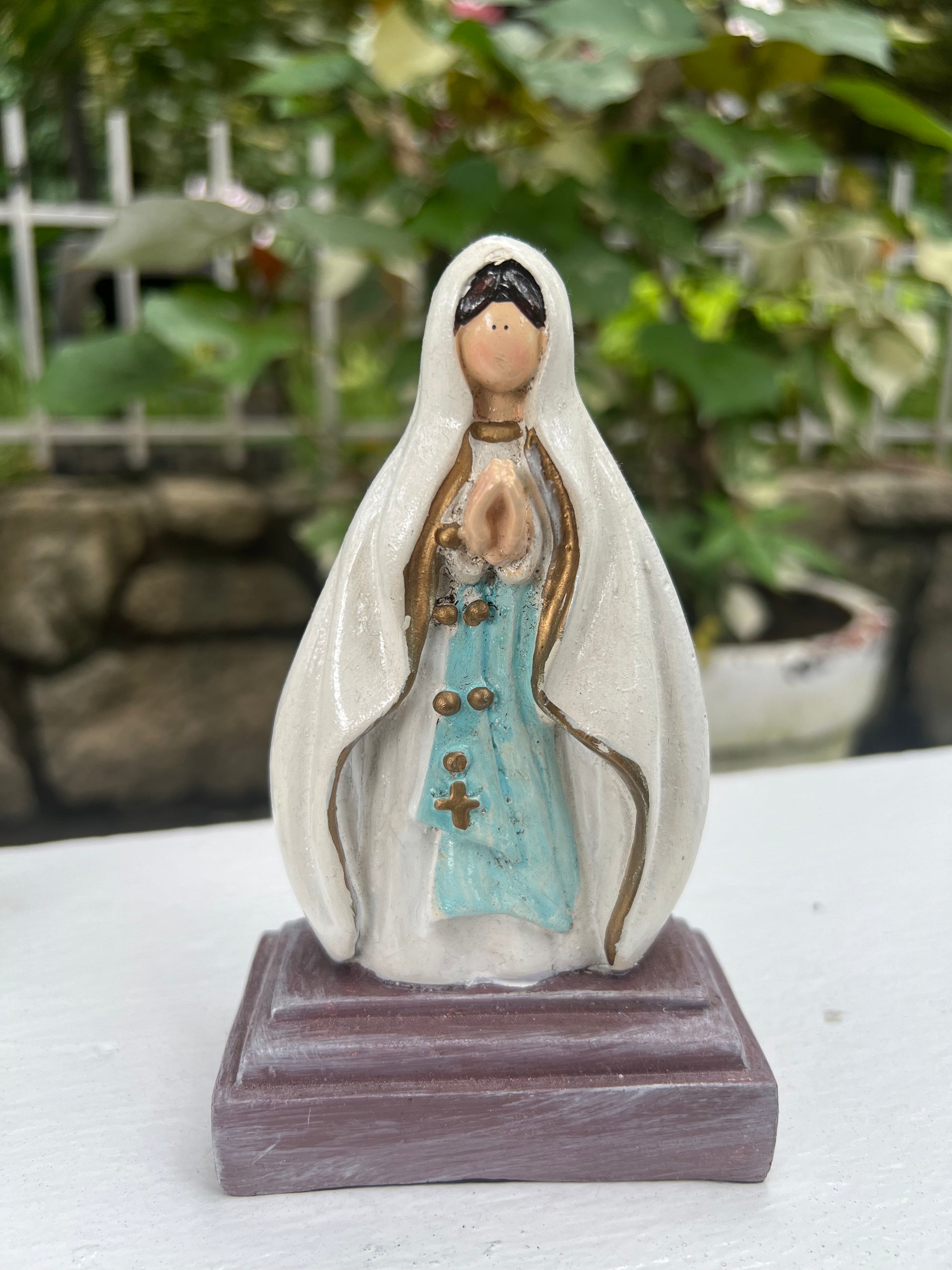 A hand-painted statue of Our Lady of Lourdes depicted with a white robe, light blue sash, and hands clasped in prayer, adorned with subtle gold details. The figurine stands on a dark brown rectangular base, set against a soft-focus background of lush green leaves and a white garden fence, suggesting an outdoor or garden setting.
