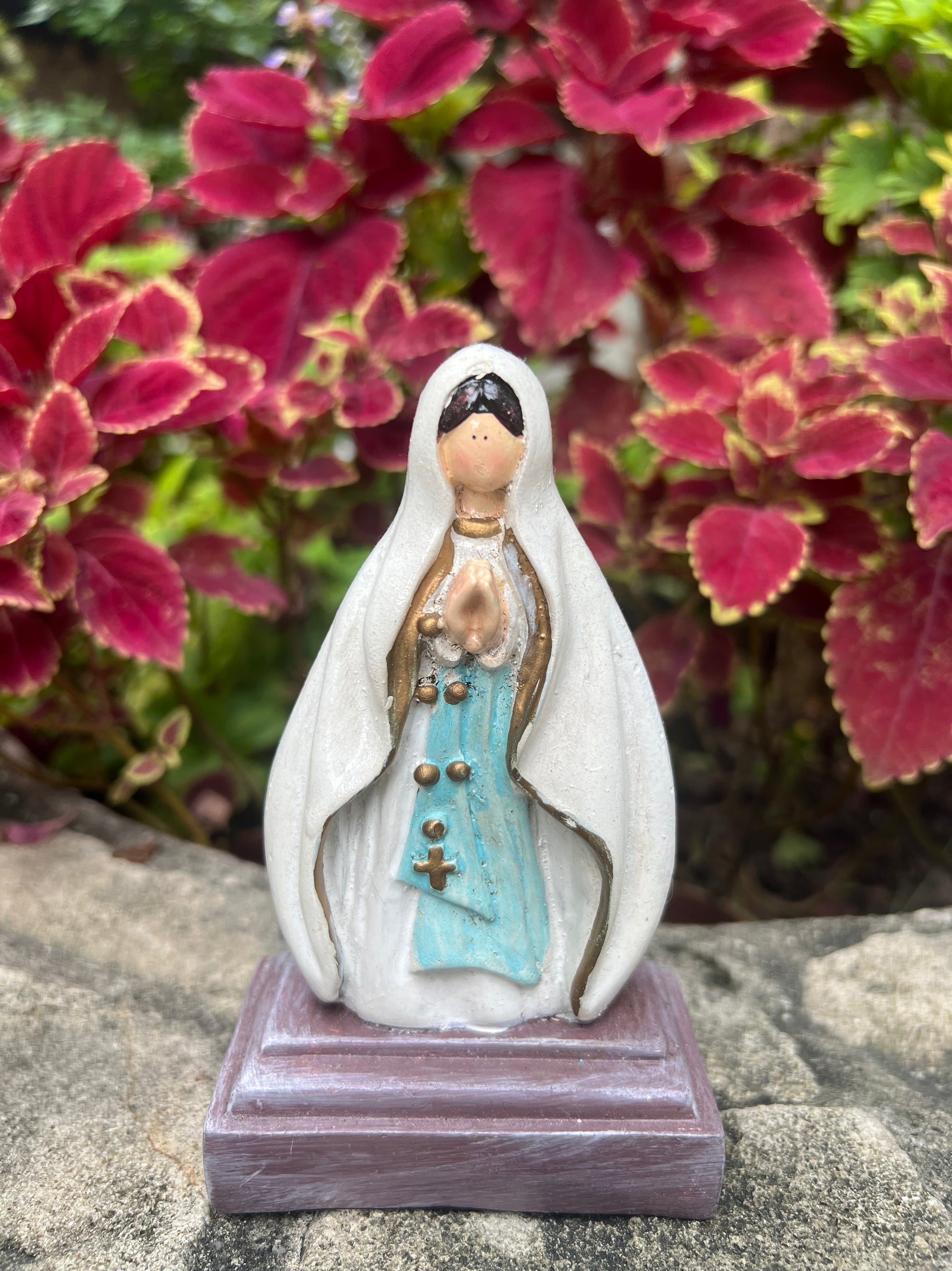 A hand-painted statue of Our Lady of Lourdes depicted with a white robe, light blue sash, and hands clasped in prayer, adorned with subtle gold details. The figurine stands on a dark brown rectangular base, set against a soft-focus background of lush red leaves, suggesting an outdoor or garden setting."
