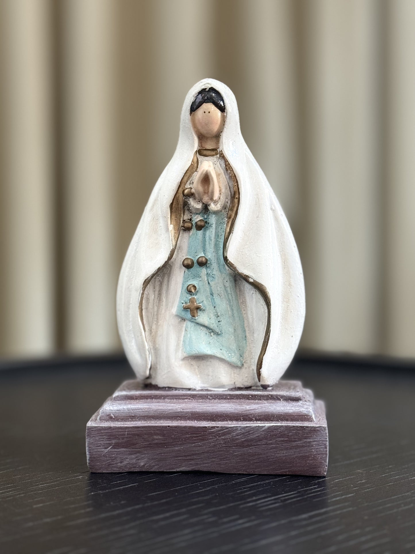 A closed-up image of a hand painted Our Lady of Lourdes statue with a white veil and light blue sash, adorned with gold accents. The statue is set against a soft neutral background .