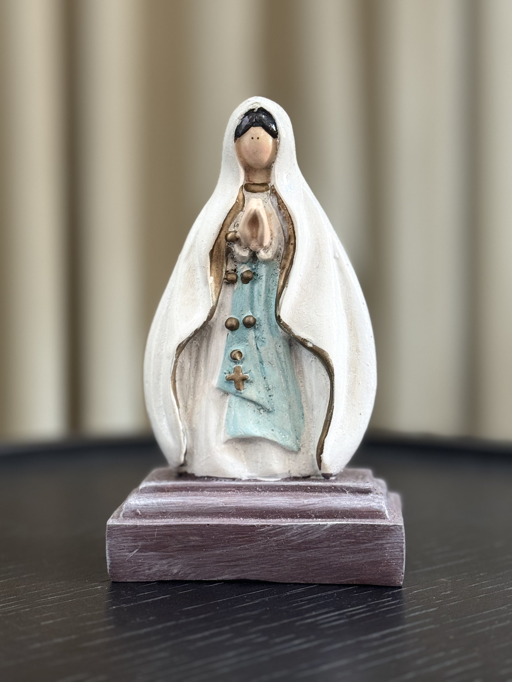 A closed-up image of a hand painted Our Lady of Lourdes statue with a white veil and light blue sash, adorned with gold accents. The statue is set against a soft neutral background .