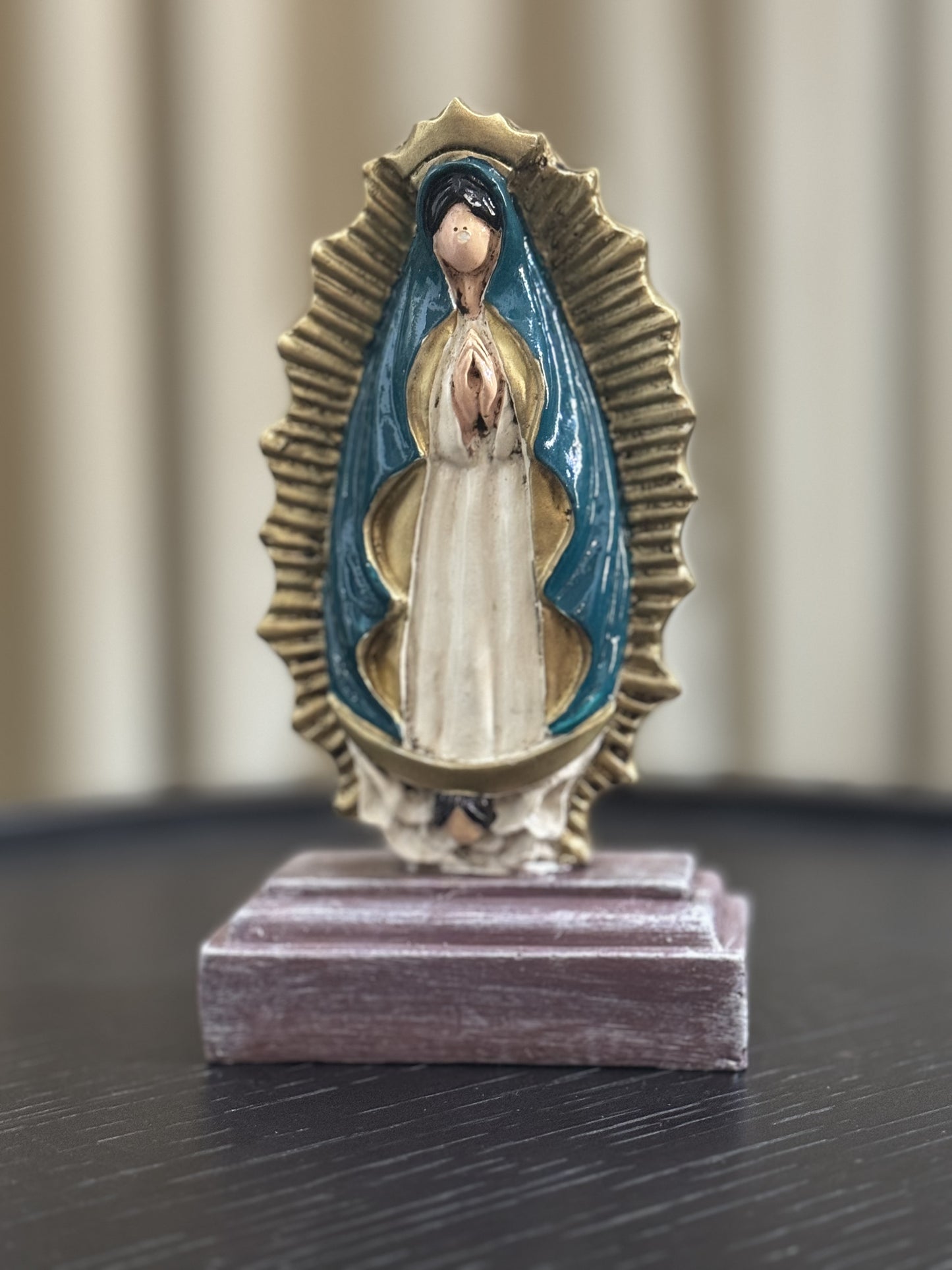 A statue of Our Lady of Guadalupe, depicted with hands clapped in a prayer, a white colored gown, dark teal cloak and framed by a radiant gold aura. The statue is place in a wooden base against a neutral indoor background. 