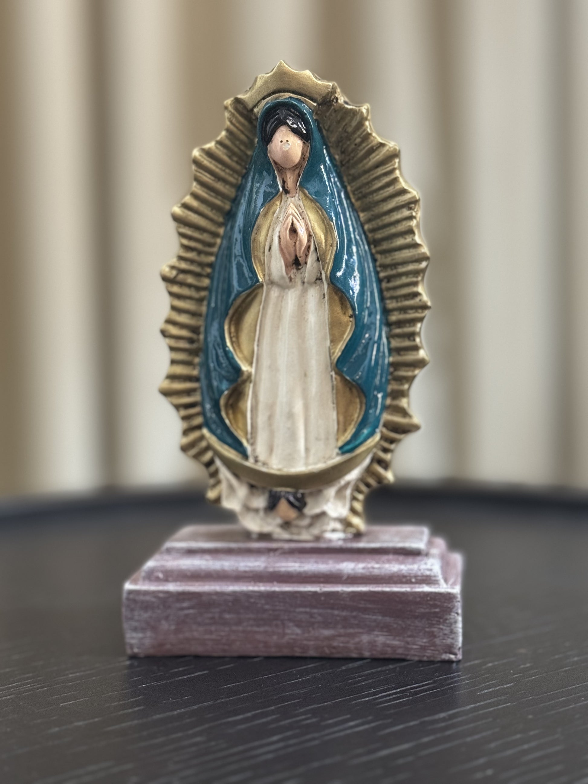 A statue of Our Lady of Guadalupe, depicted with hands clapped in a prayer, a white colored gown, dark teal cloak and framed by a radiant gold aura. The statue is place in a wooden base against a neutral indoor background. 