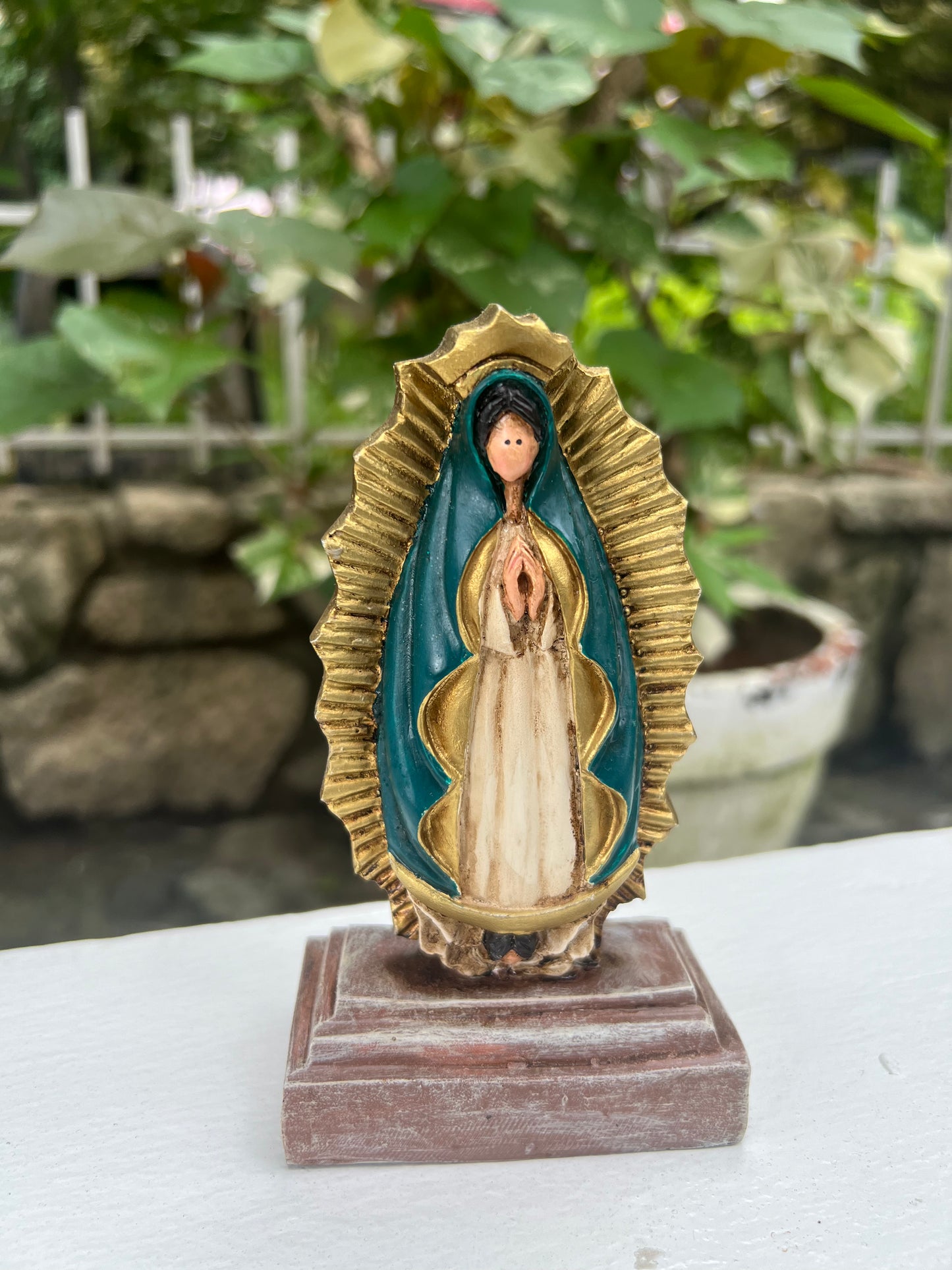 A statue of Our Lady of Guadalupe, depicted with hands clapped in a prayer, a white colored gown, dark teal cloak and framed by a radiant gold aura. The statue is place in a wooden base against a garden background. 