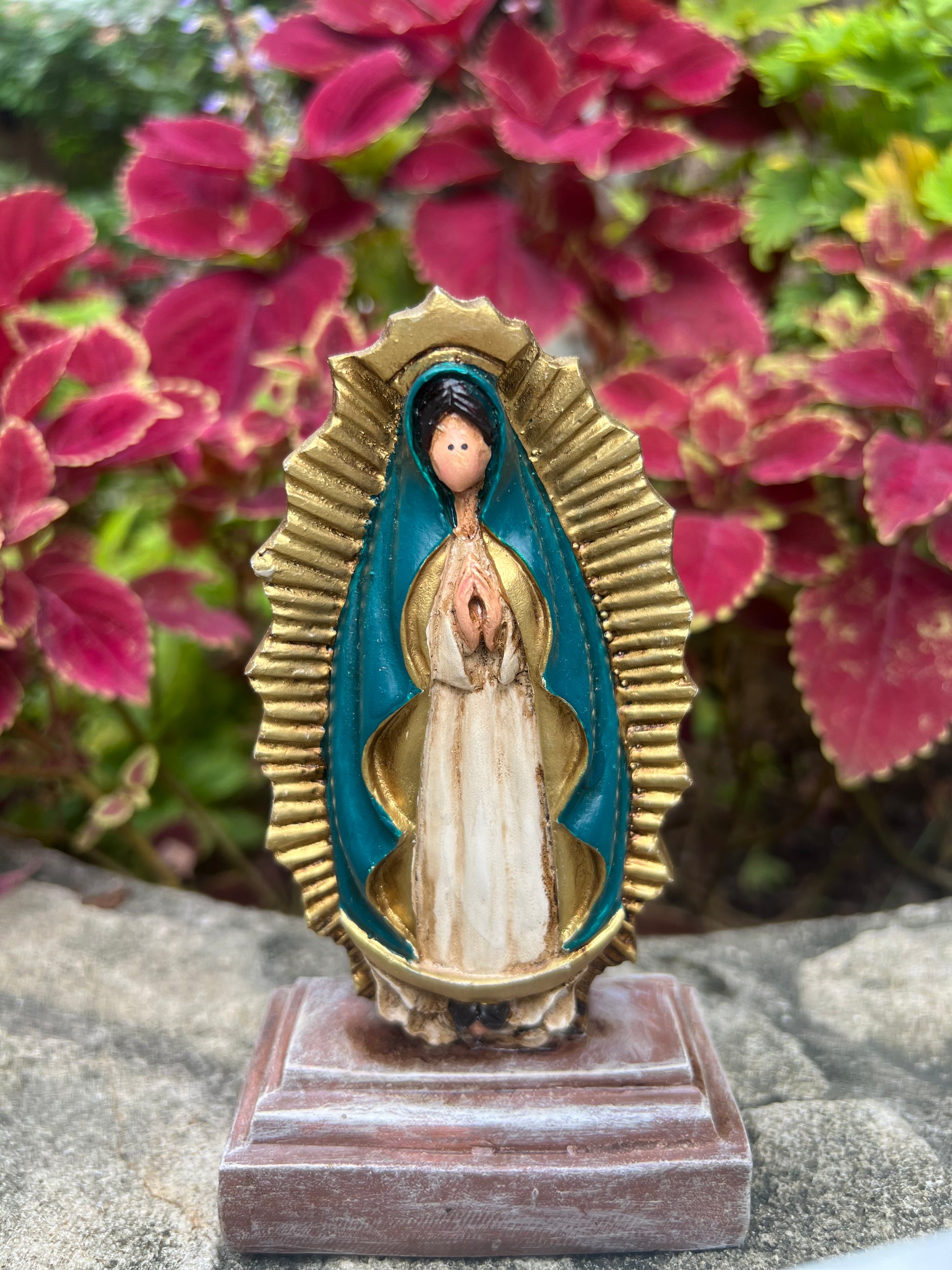 A statue of Our Lady of Guadalupe, depicted with hands clapped in a prayer, a white colored gown, dark teal cloak and framed by a radiant gold aura. The statue is place in a wooden base against a garden background.