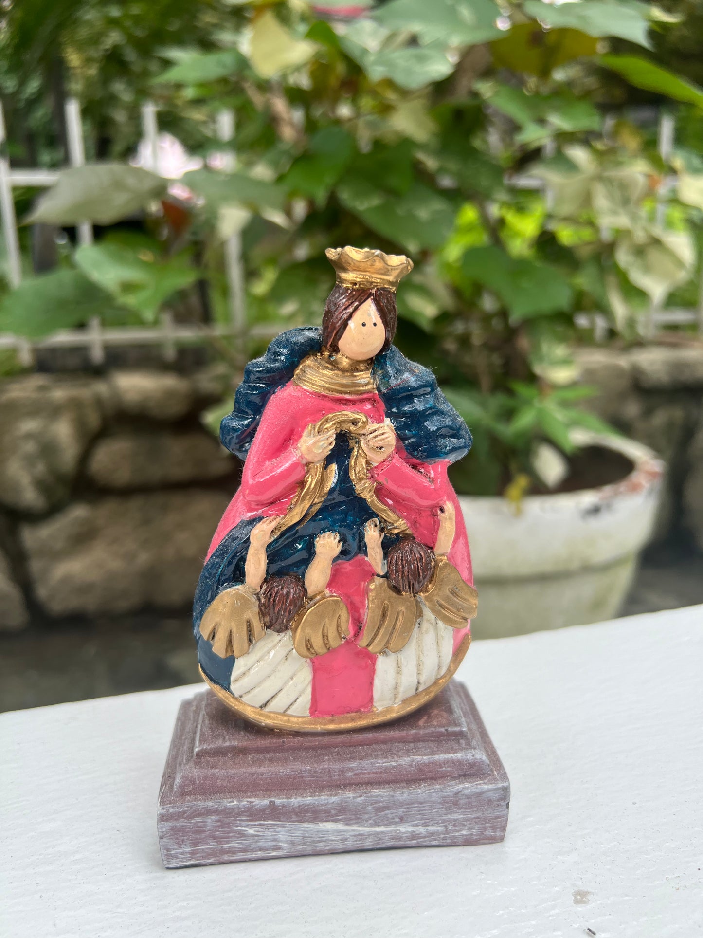 A hand-painted statue of Our Lady, Undoer of Knots, depicting Mary in a pink and blue, crowned, and surrounded by angels, untying a knotted ribbon. The statue is set outdoors with blurred green foliage in the background.