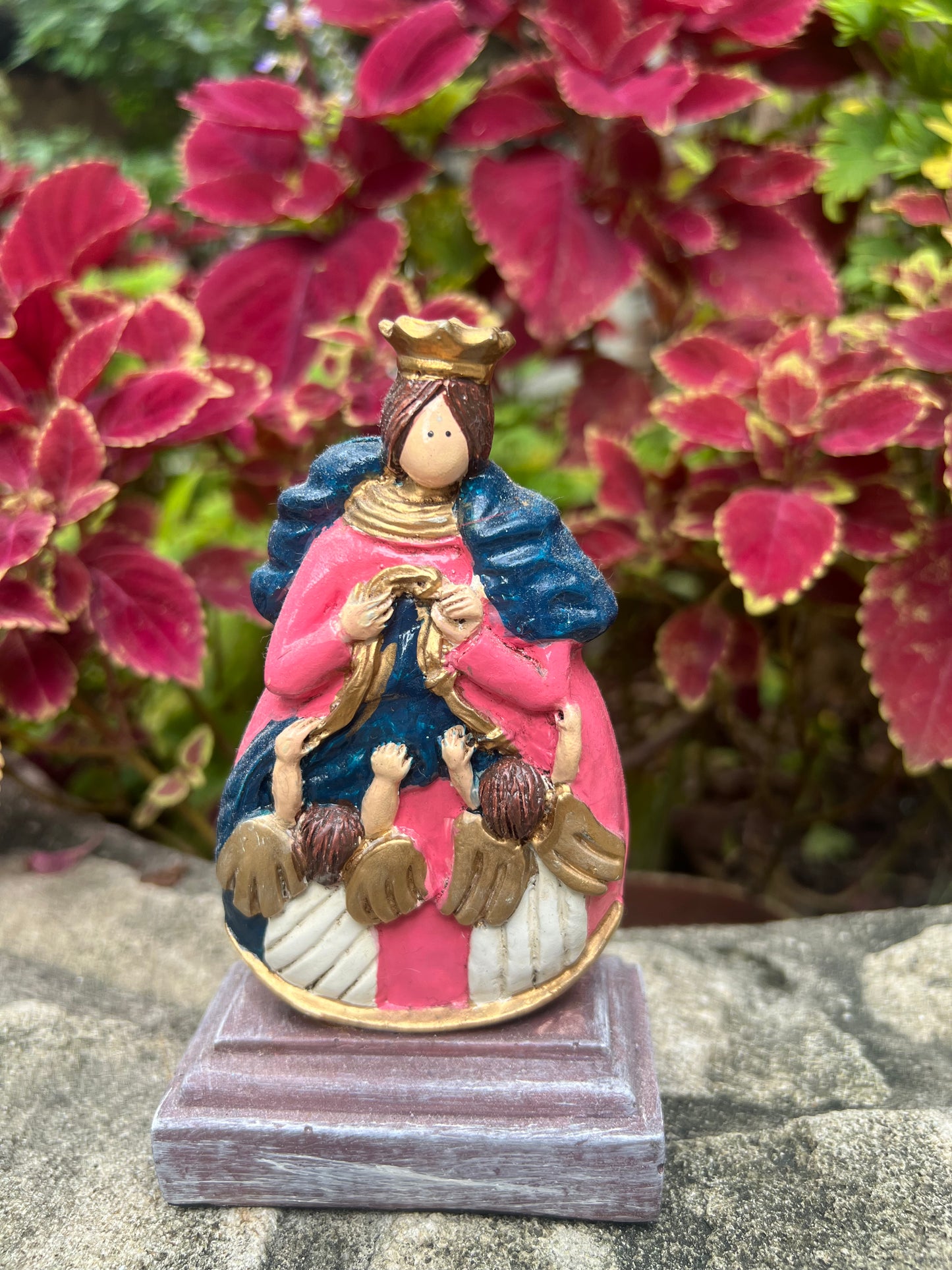 A colorful, hand-painted statue of Our Lady, Undoer of Knots, depicted with a gold crown, pink and blue robes, and surrounded by angels untying knots. The statue is displayed outdoors against a vibrant backdrop of red and green leaves.