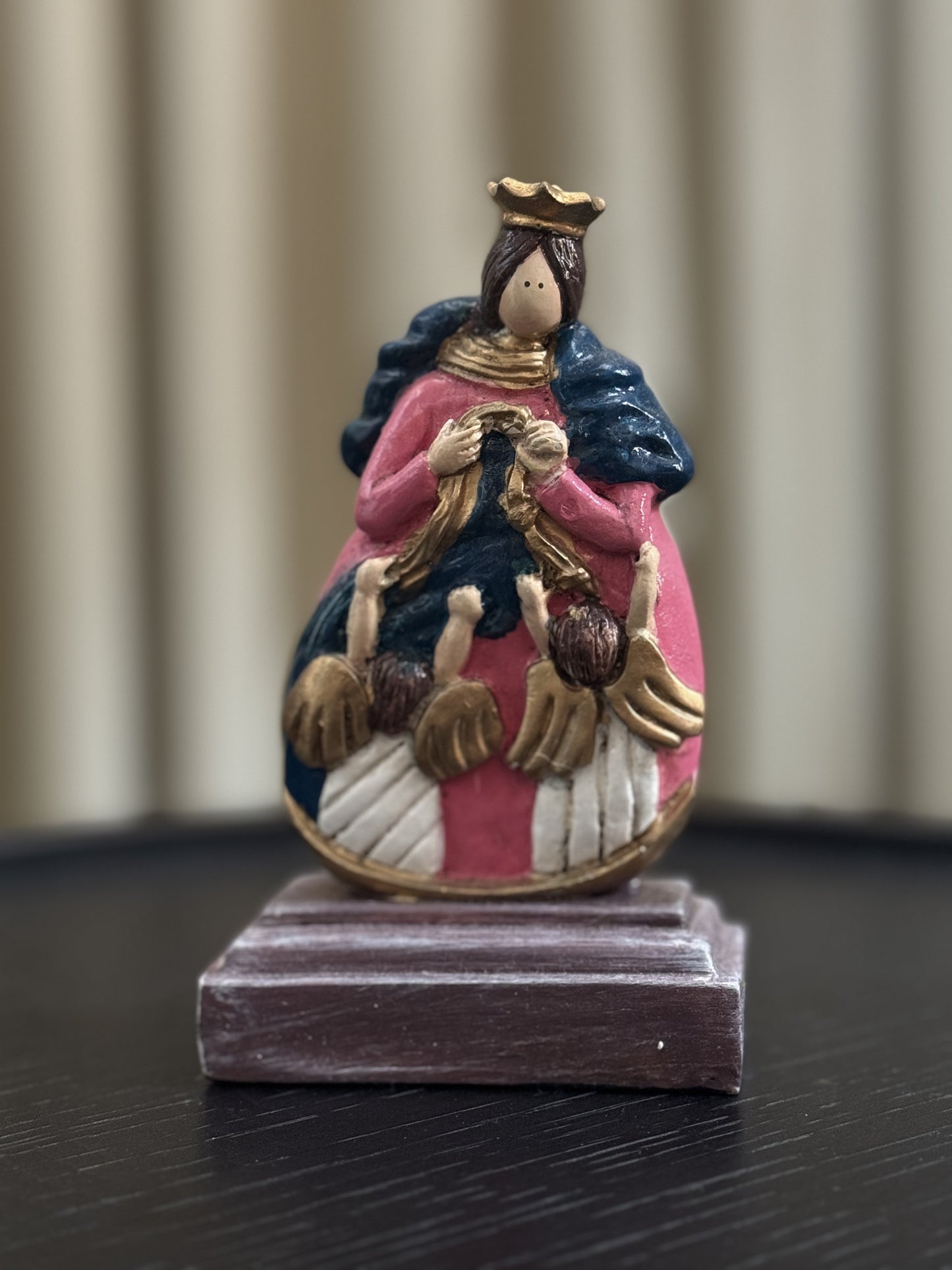 A colorful hand-painted figurine of Our Lady, Undoer of Knots, depicting Mary in a golden crown, pink and blue, untying a knotted ribbon, surrounded by angels. The statue rests on a dark wooden base against a blurred, neutral indoor background.