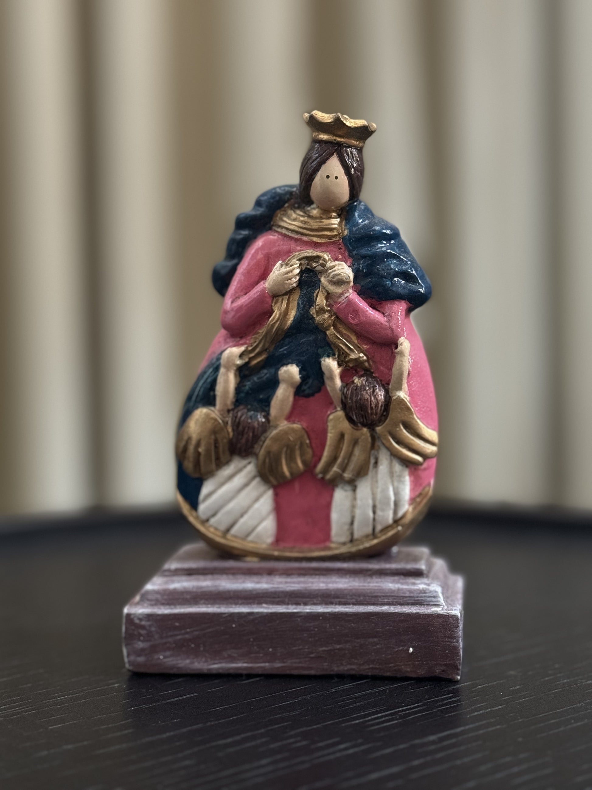 A colorful hand-painted figurine of Our Lady, Undoer of Knots, depicting Mary in a golden crown, pink and blue, untying a knotted ribbon, surrounded by angels. The statue rests on a dark wooden base against a blurred, neutral indoor background.
