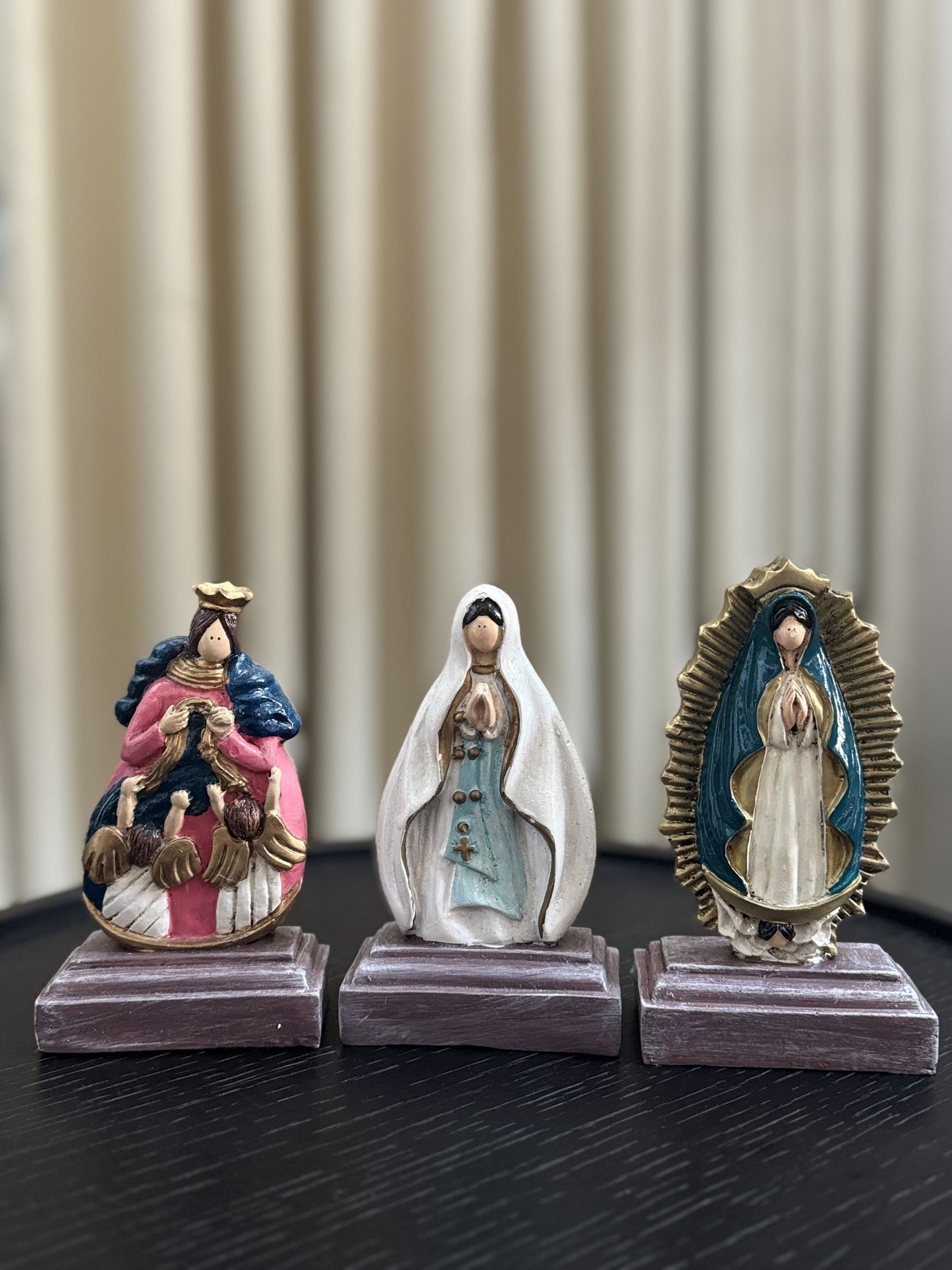 A display of three statue of the Virgin Mary, each with unique iconography and vibrant colors. From left to right: Our Lady, Undoer of Knots, adorned in a pink and blue robe with angels; Our Lady of Lourdes in a flowing white robe and blue sash; and Our Lady of Guadalupe framed by a radiant golden aura. Each figurine is placed on a simple, square wooden base, suggesting a personal altar or collection.