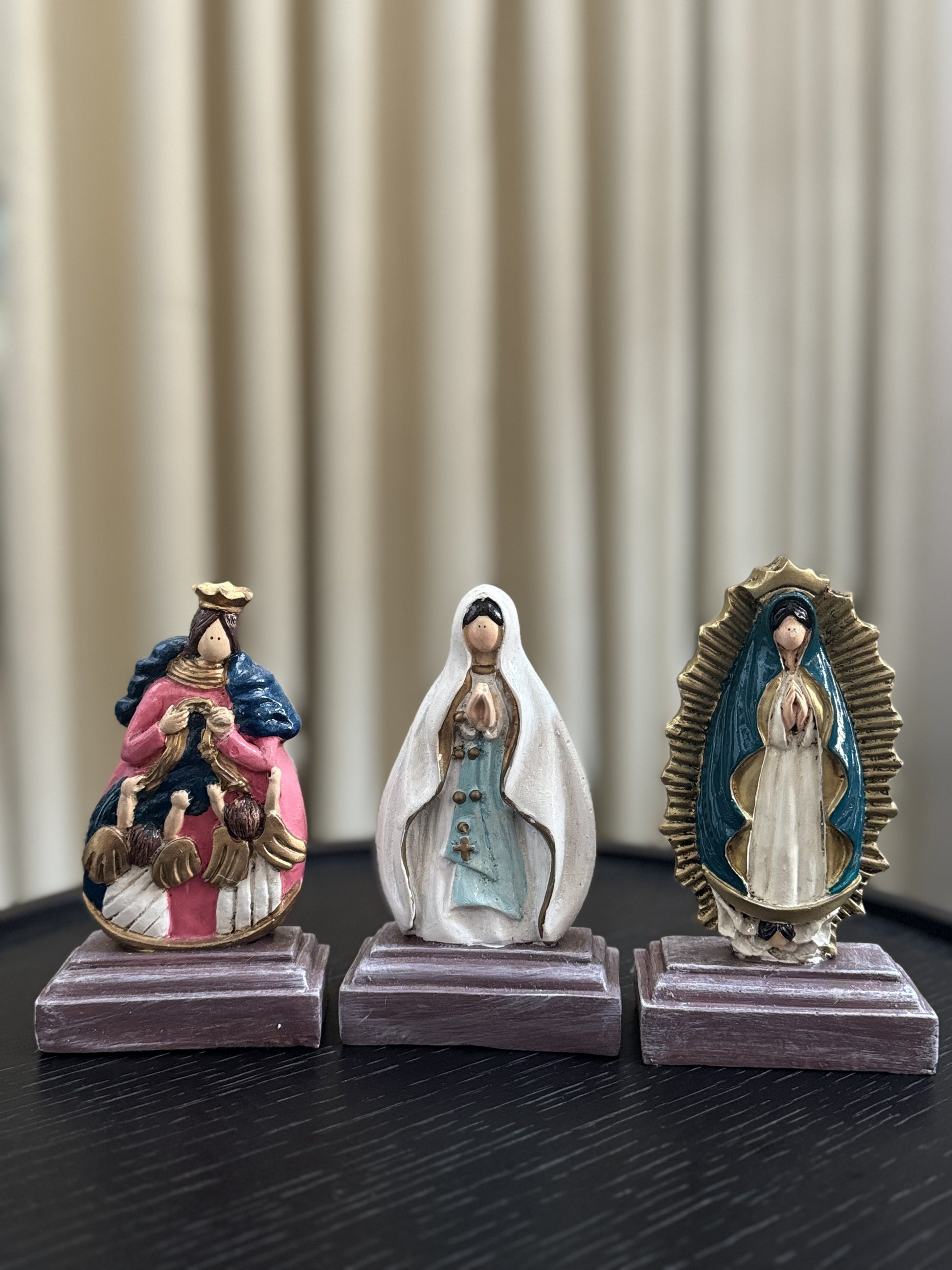 A display of three statue of the Virgin Mary, each with unique iconography and vibrant colors. From left to right: Our Lady, Undoer of Knots, adorned in a pink and blue robe with angels; Our Lady of Lourdes in a flowing white robe and blue sash; and Our Lady of Guadalupe framed by a radiant golden aura. Each figurine is placed on a simple, square wooden base, suggesting a personal altar or collection.