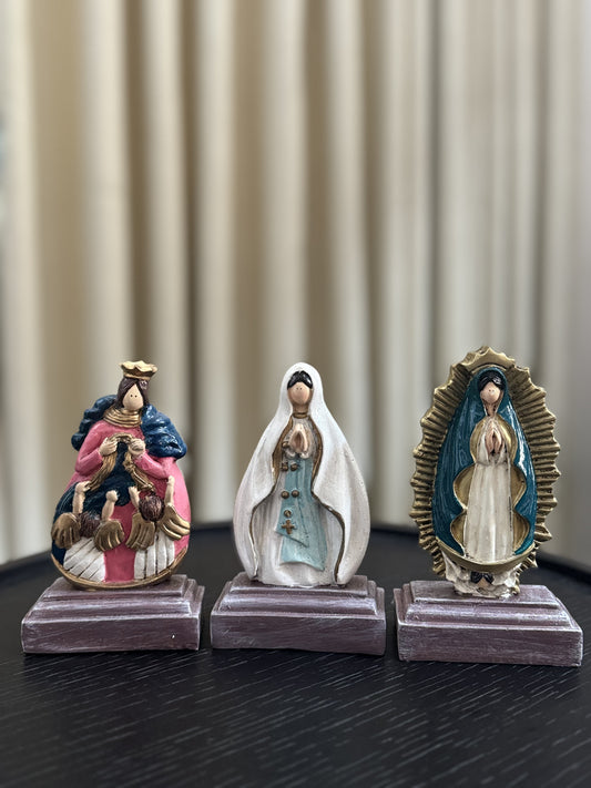 A display of three statue of the Virgin Mary, each with unique iconography and vibrant colors. From left to right: Our Lady, Undoer of Knots, adorned in a pink and blue robe with angels; Our Lady of Lourdes in a flowing white robe and blue sash; and Our Lady of Guadalupe framed by a radiant golden aura. Each figurine is placed on a simple, square wooden base, suggesting a personal altar or collection.